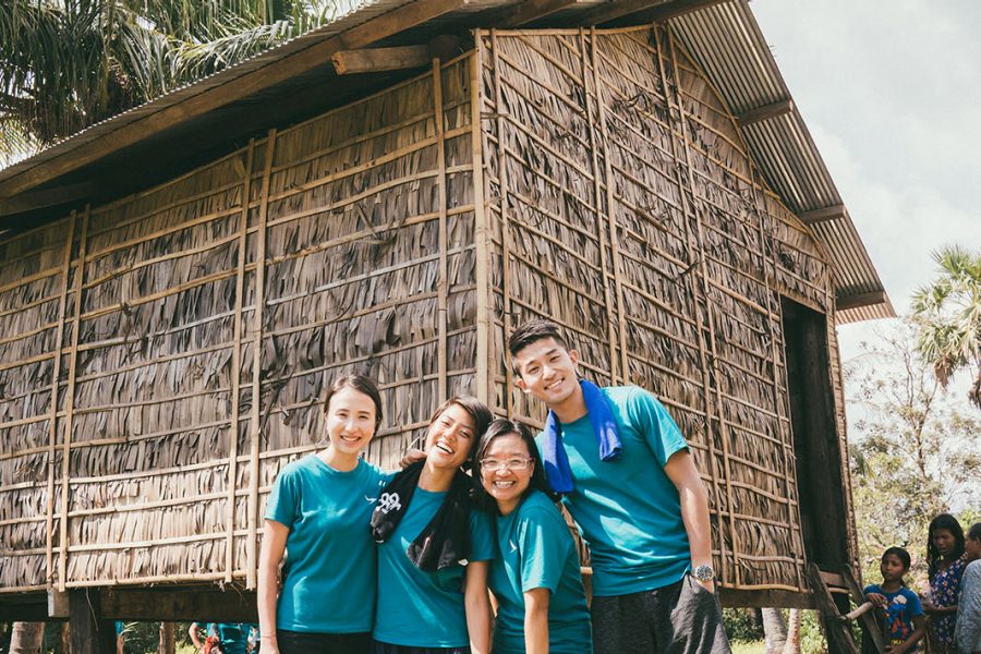 Four people in matching blue shirts stand in front of a traditional bamboo and thatch stilt house, with palm trees and a partly cloudy sky in the background.