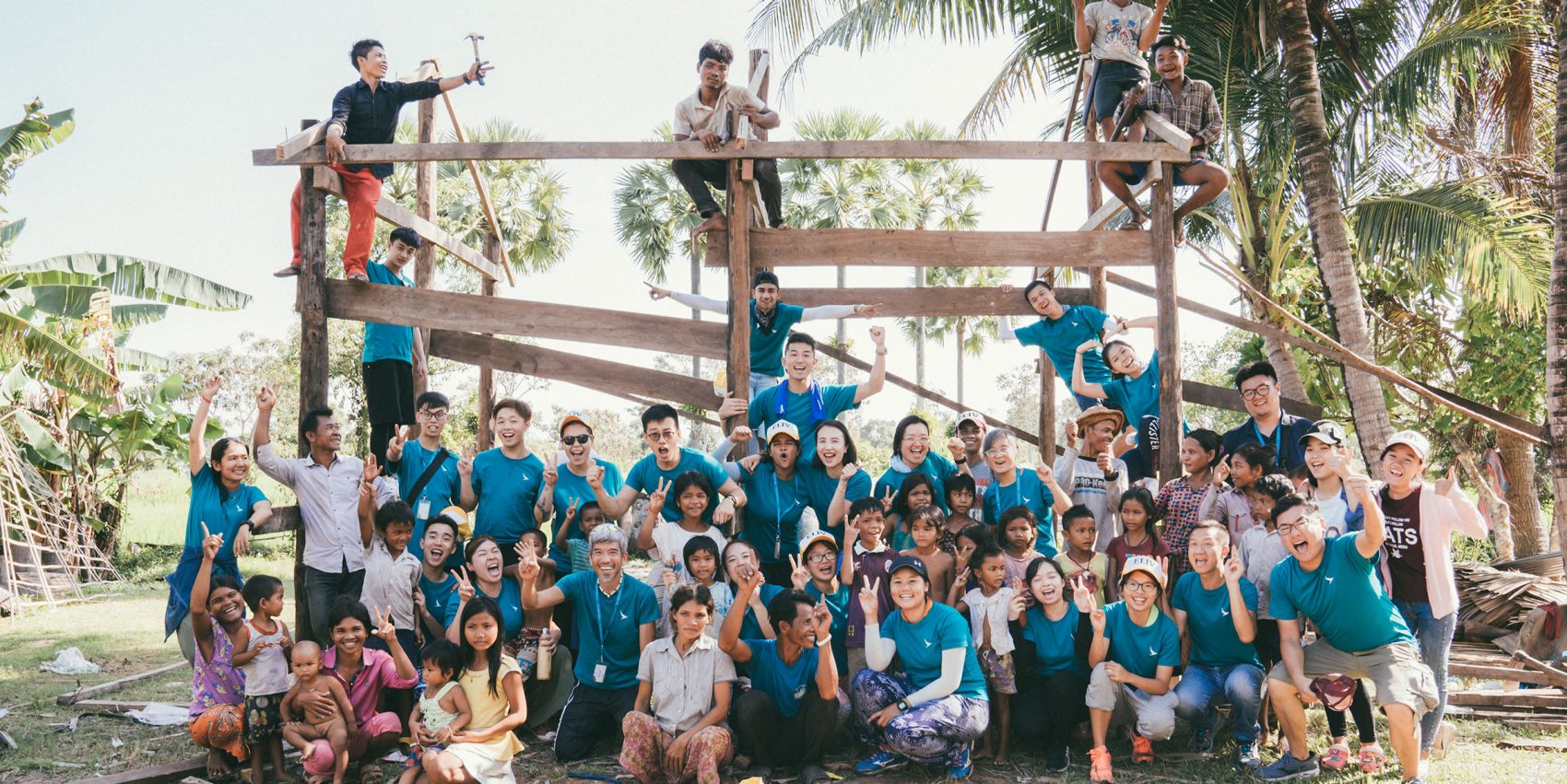A large group of people, including adults and children, gathered in front of a wooden structure under construction, with some standing on it holding tools, suggesting a community building project amid palm trees and greenery.