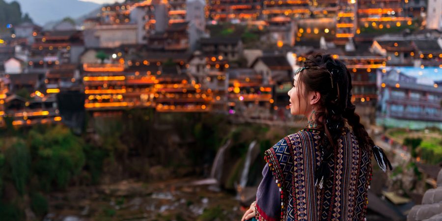 一位身穿部落服飾的女性旅客，站在觀景台上俯瞰中�國內地的一座古老城鎮。