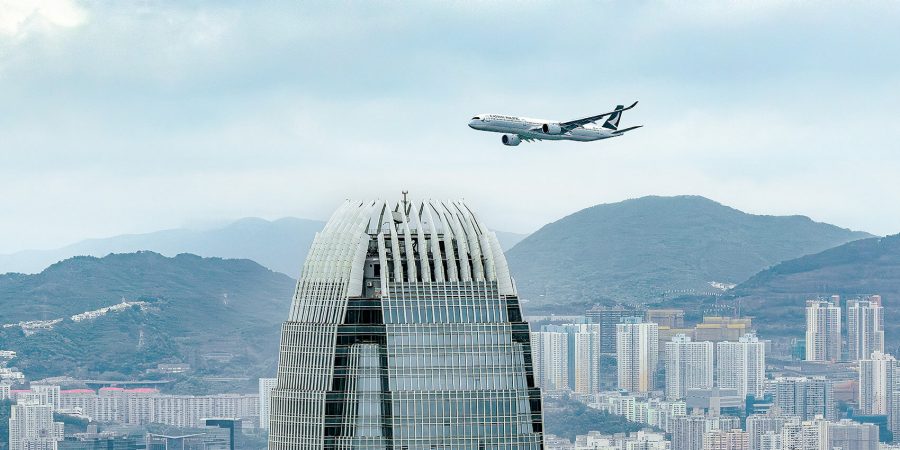 Cathay Pacific airplane flying above a tall modern skyscraper with a curved top, set against a cityscape of high-rise buildings and distant green mountains under a cloudy sky.