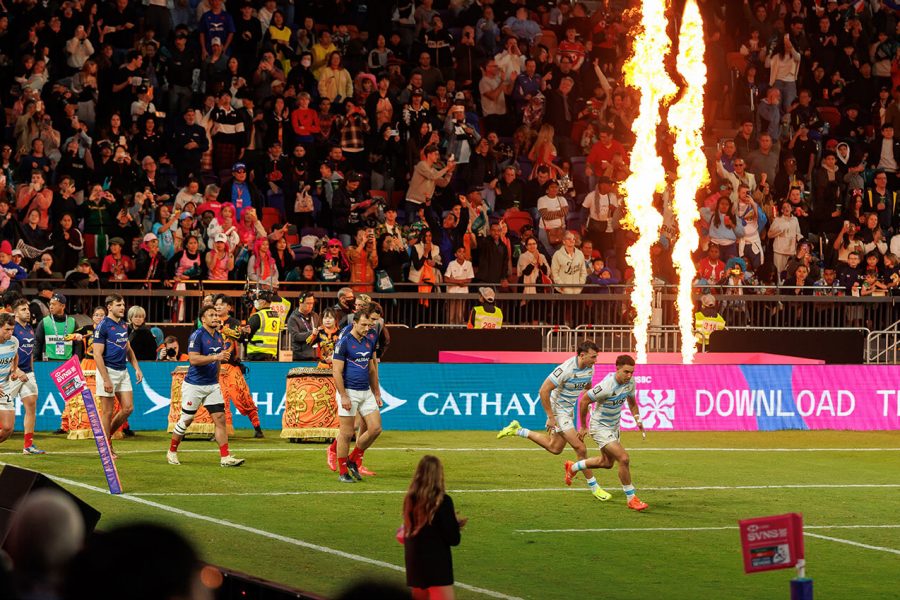 Rugby players running on a brightly lit stadium field during a match, with flames shooting up as part of the event spectacle. A large crowd fills the stands in the background, and colorful advertising boards display messages including ‘Cathay’ and ‘Download the HSBC.