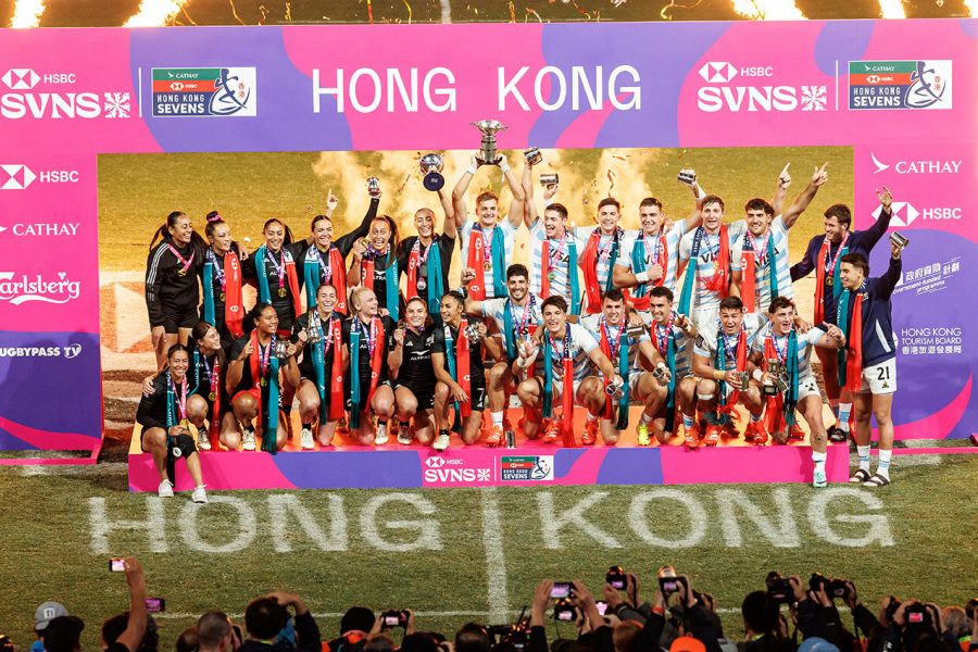 Rugby team celebrating on a podium at the Hong Kong Sevens tournament, holding trophies and wearing medals, with a large pink and purple backdrop displaying ‘HONG KONG’ and sponsor logos including HSBC, SVNS, Cathay, and Carlsberg. Bright flames shoot upward on both sides, and spectators in the foreground capture the moment with cameras.