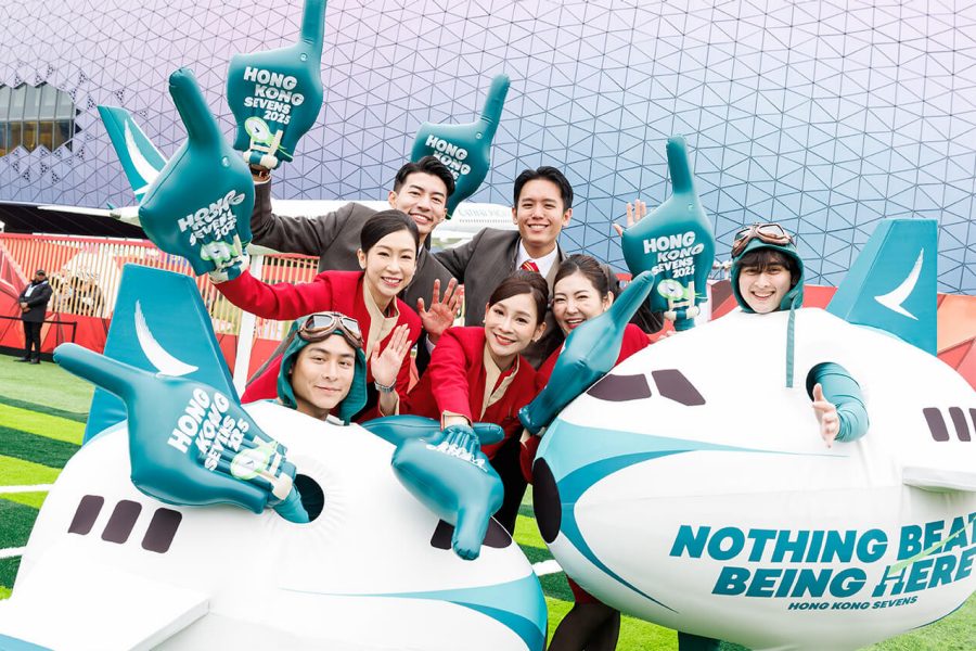 Group of people posing outdoors on a green sports-themed surface, holding large foam hands with the text ‘HONG KONG SEVENS,’ and sitting inside inflatable airplane props branded with Cathay Pacific and the slogan ‘NOTHING BEATS BEING HERE.’ A modern building with a geometric patterned facade and a red barrier is visible in the background.