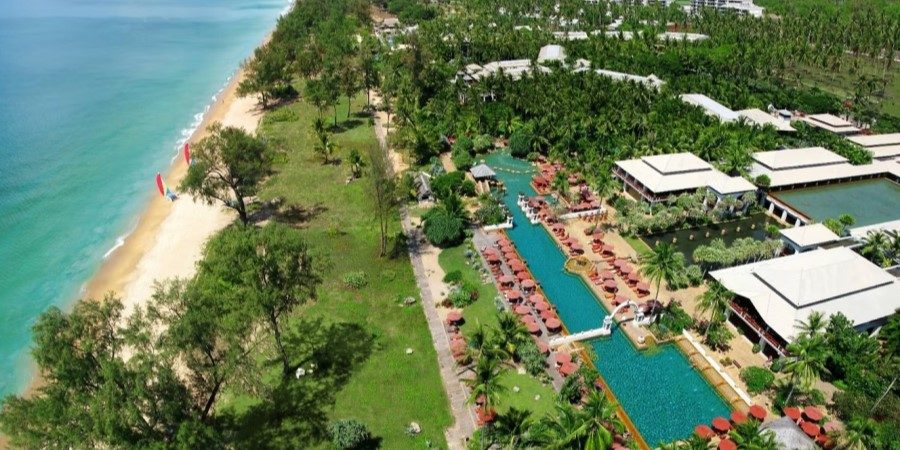 Aerial view of a beach resort, showcasing sandy shores, turquoise waters, and palm trees under a clear blue sky.