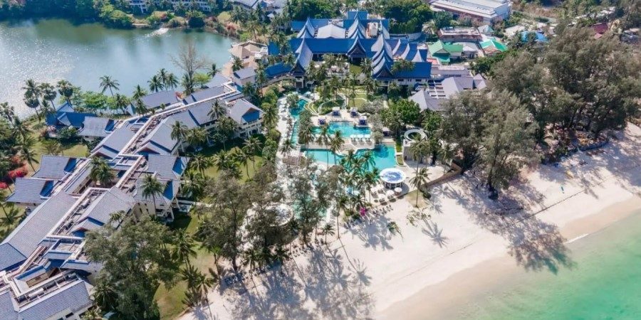 Aerial view of a resort with lush greenery and a sandy beach, showcasing the ocean waves and sunbathers below.