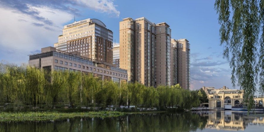 A river flows through a cityscape, flanked by tall buildings and lush trees on either side.