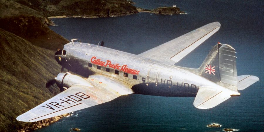 Un appareil vintage de Cathay Pacific survolant une côte escarpée, avec l’inscription « Cathay Pacific Airways » en rouge sur le fuselage en métal nu. 
