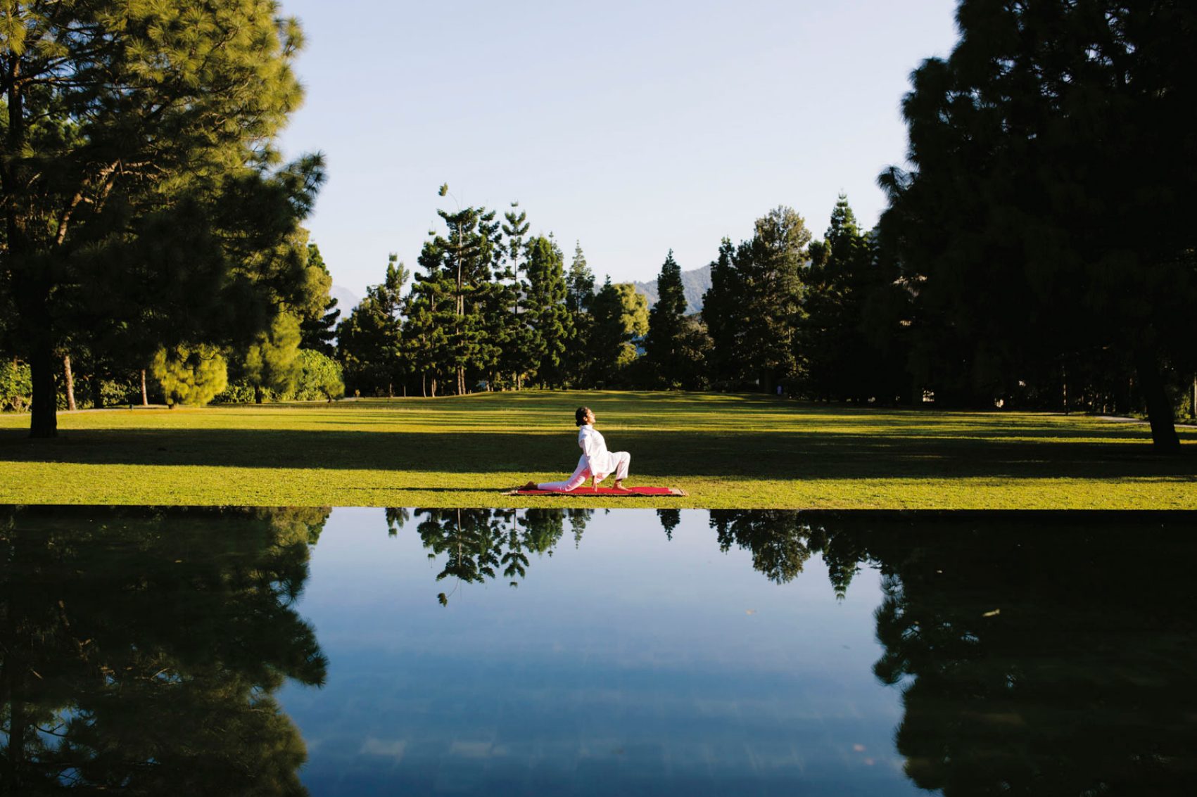 A woman doing yoga on a mat in the middle of a garden infront of a pond surrounded by green grass and trees in Ananda Uttarakhand, India