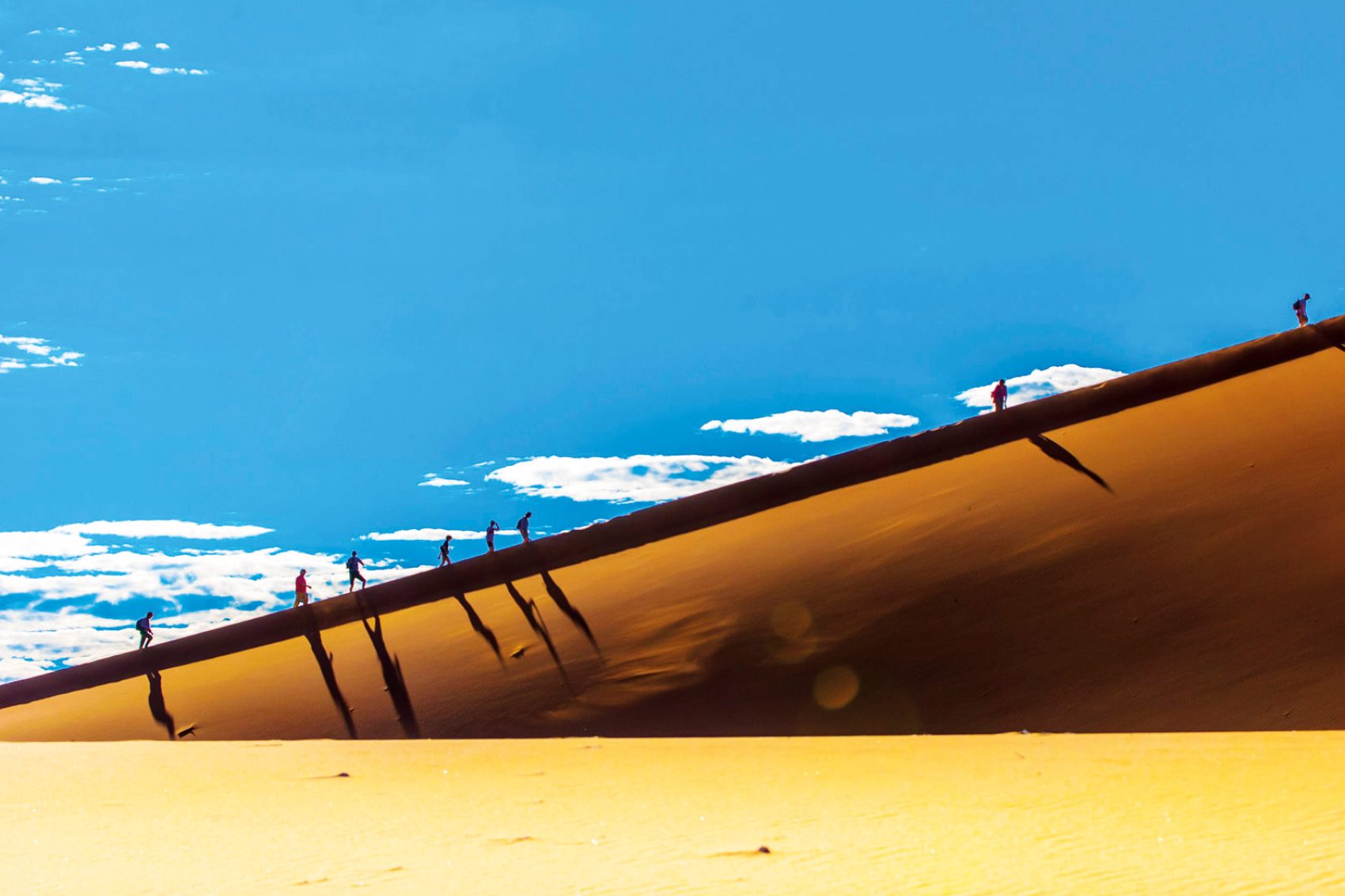 People climbing up a steep sandune with a blue sky backdrop