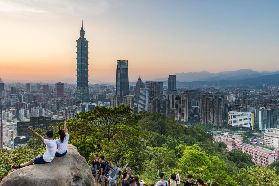 Two people sitting on a rock holding peace signs in the air, looking out at a view of Taipei 101 and other skyscrapers in the distance.