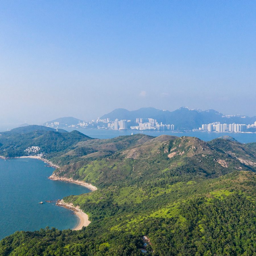 An aerial view of rolling green hills surrounded by blue seas in Mount Stenhouse and Ling Kok Shan, Lamma Island with Hong Kong's skyline and skyscrappers in the background