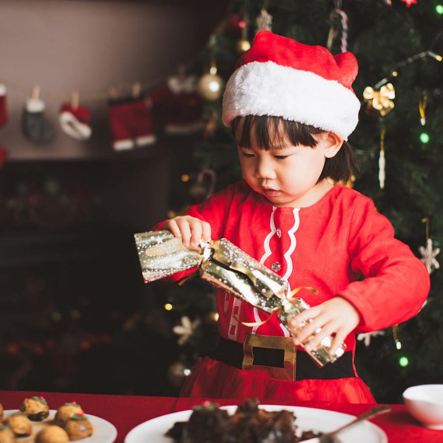 toddler girl in Santa outfit playing with a Christmas cracker in front of Christmas tree 