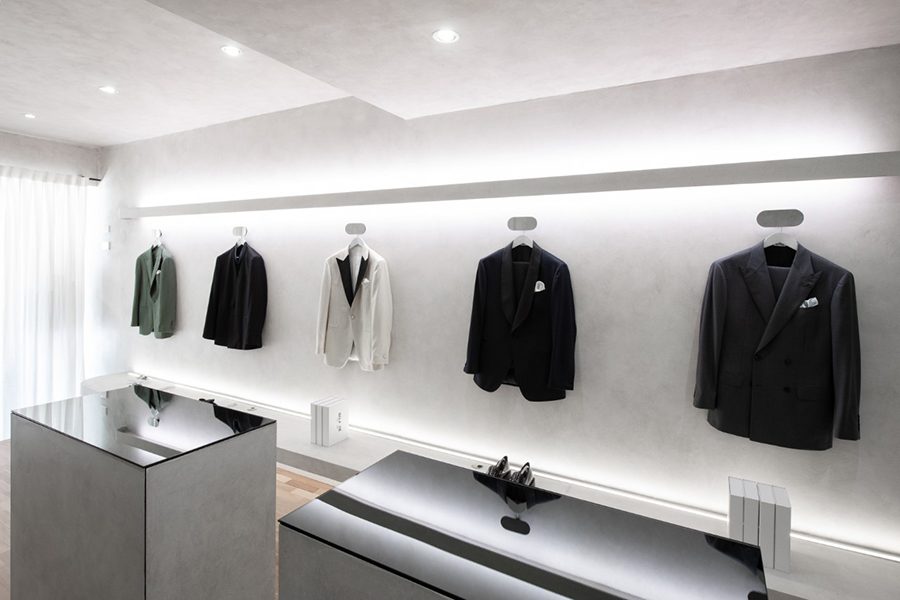 An interior view of Belance showroom featuring various styles of dress shoes and bespoke suits hanging on the display wall