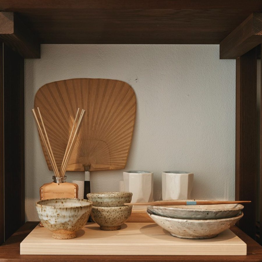 A close up view of ceramic bowls, cups, plates, wooden chopsticks, paper fan, fragrance and home accessores on display on a wooden shelf