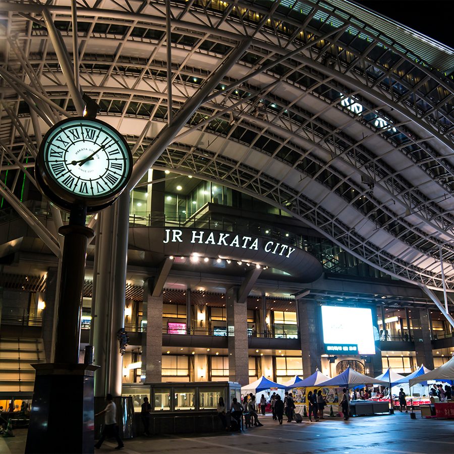 An exterior view of JR Hakata City Amu Plaza entrance at night with pop up stalls and a clock tour outside