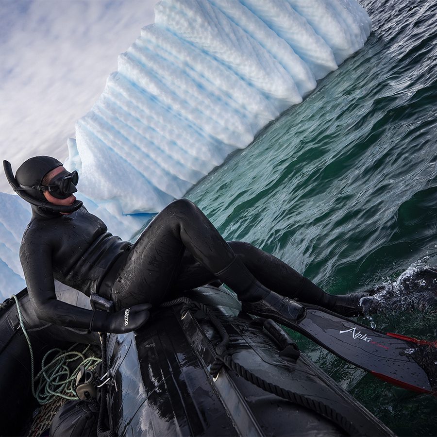 A man in a black wet suit and snorkling goggles climbing out of a black inflatable boat to ice dive in Antartica