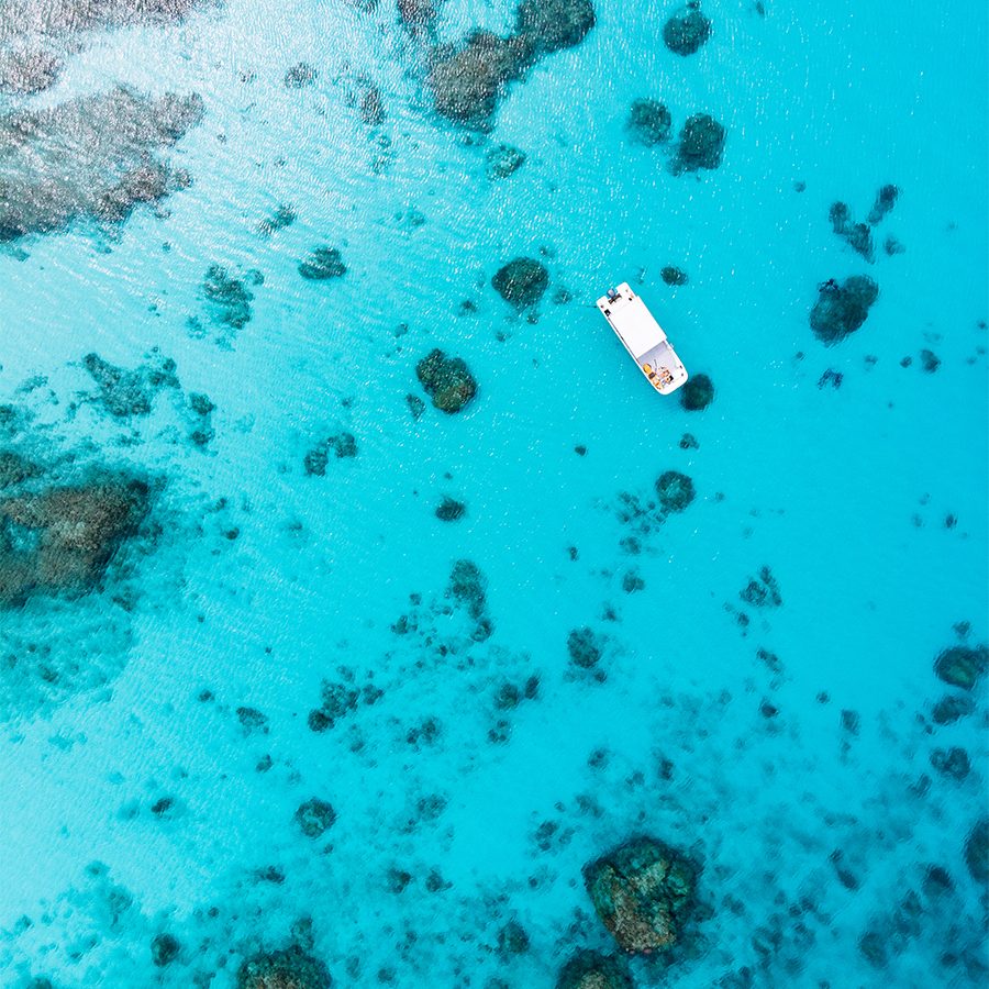 A top down aerial view of a boat in the open sea surrounded by blue water and corals in the ocean in Okinawa, Japan