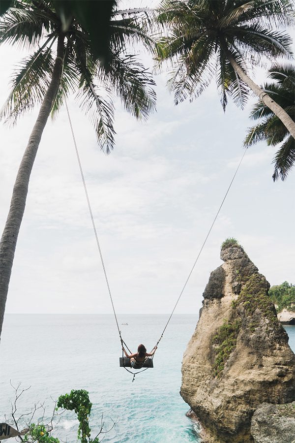 A person sitting on a swing between two palm trees overlooking the ocean