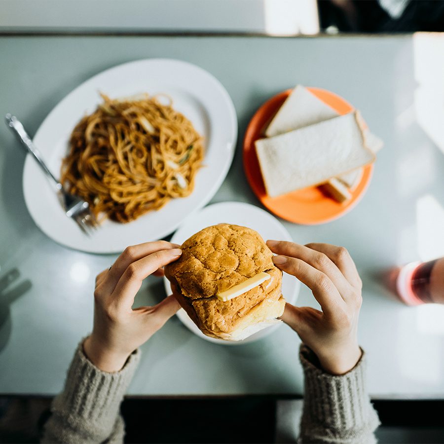A person holding a pineapple bun and  butter with a plate of noodles and toast on the table
