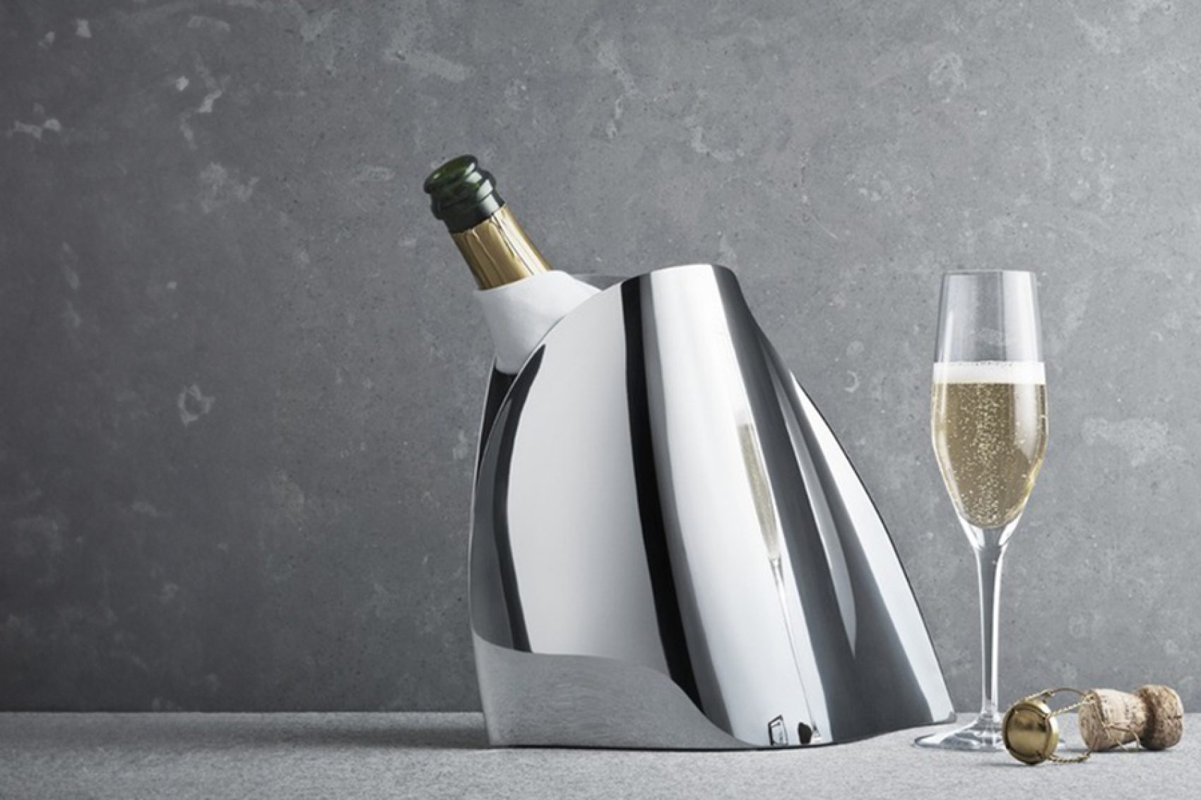 An opened bottle of champagne in a Georg Jensen stainless steel champagne cooler bucket next to a glass of champagne