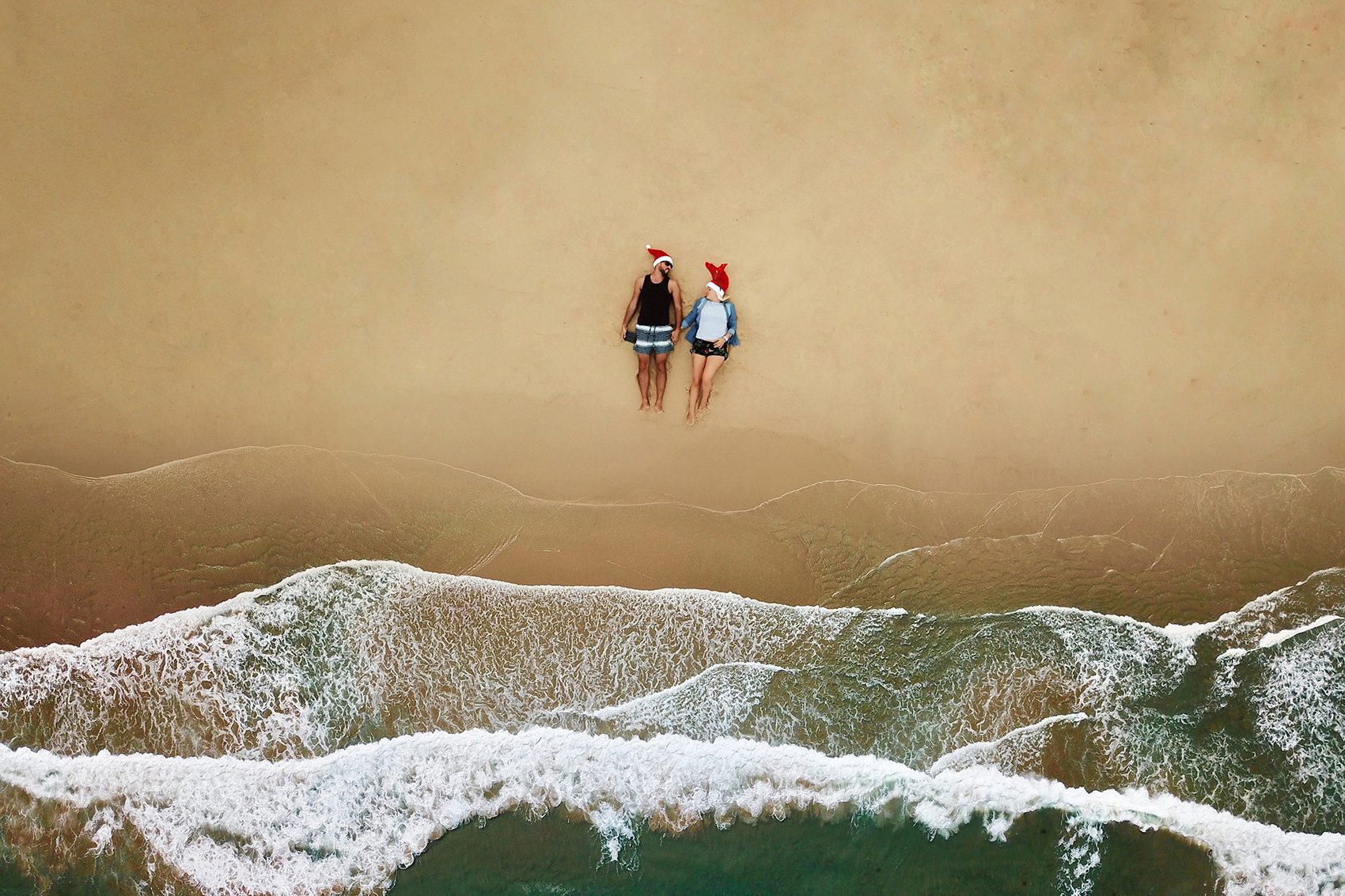 Two people lying on beach together wearing santa hats