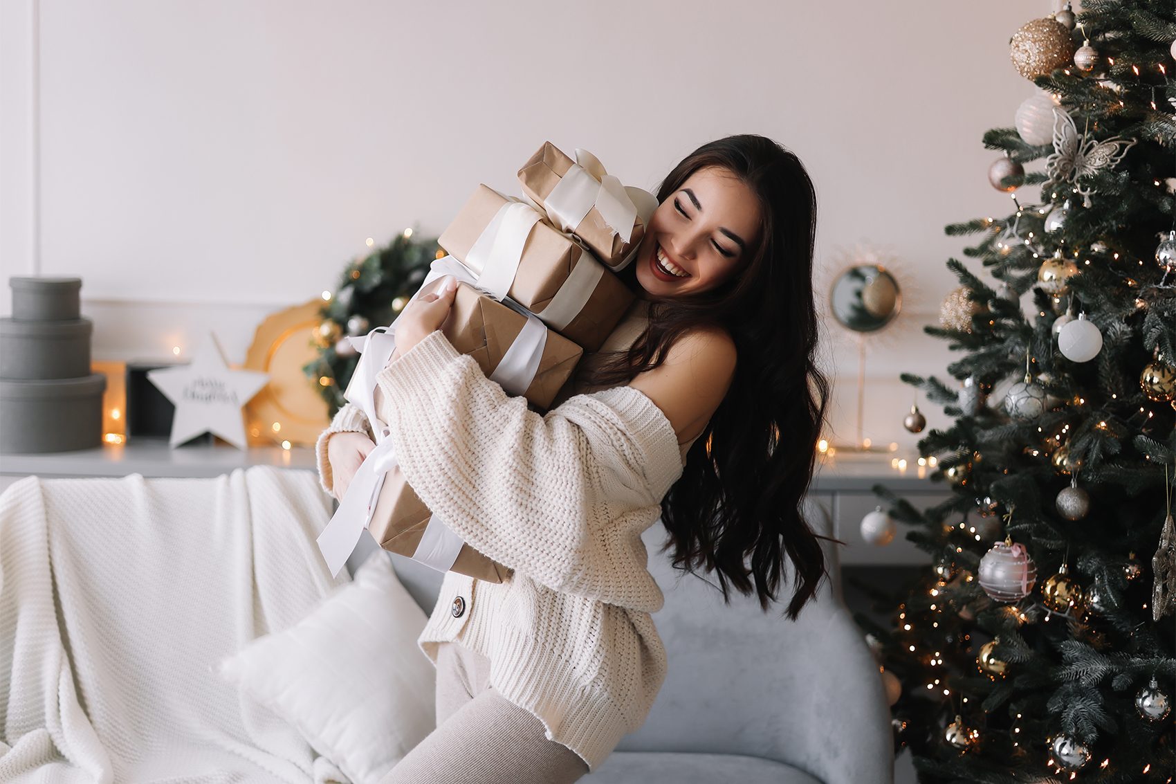 An Asian woman wearing a white sweater happily carrying a stack of wrapped Christmas presents next to a Christmas tree at home