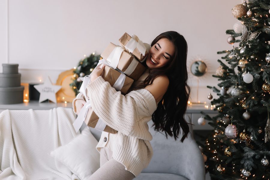An Asian woman wearing a white sweater happily carrying a stack of wrapped Christmas presents next to a Christmas tree at home