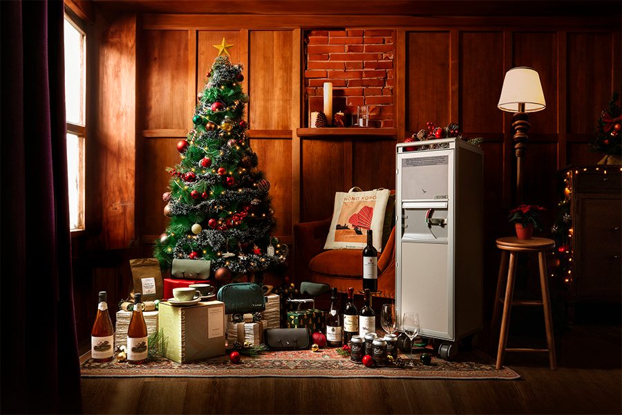 A wide shot of Cathay's exclusive holiday A350 bar cart set layed out under a Christmas tree at home, featuring premium teas, coffees, snacks, fine wines and luxury travel accessories