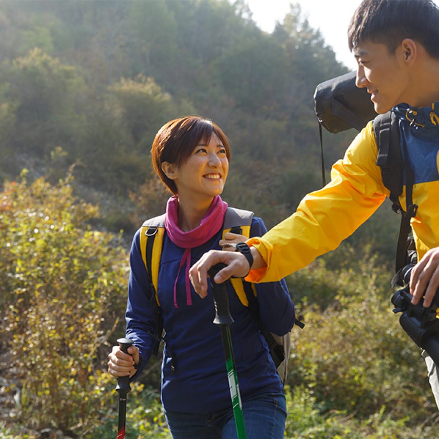 A couple of man and woman wearing hiking gear looking at each other and smiling while holding walking sticks and a pair of binoculars a sunny day