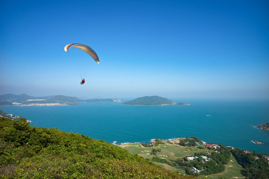 A landscape view of a paraglider flying over green hills and a golf course by the ocean on a sunny day
