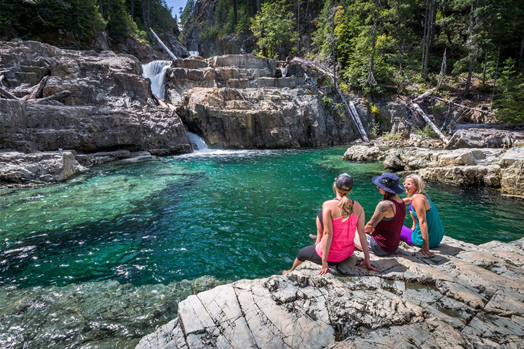 Three women sitting on a rock talking and looking at a running stream in Lower Myra Falls in Vancouver, Canada