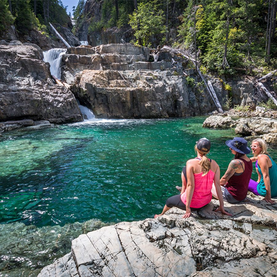 Three women sitting on a rock talking and looking at a running stream in Lower Myra Falls in Vancouver, Canada