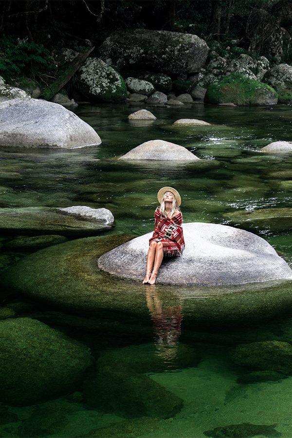 Woman in red dress and hat sits on a rock in Mossman River at Silky Oaks Lodge, Australia