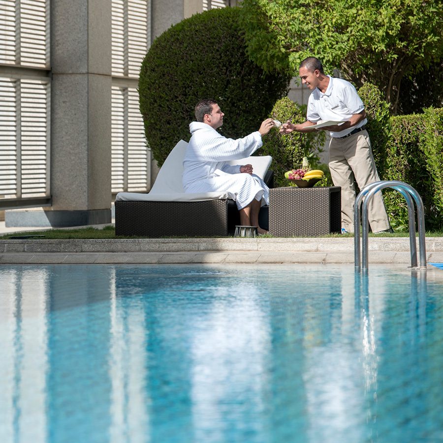 A woman sitting in the lounge area at the pool of Four Seasons Riyadh receives a drink from the waiter.