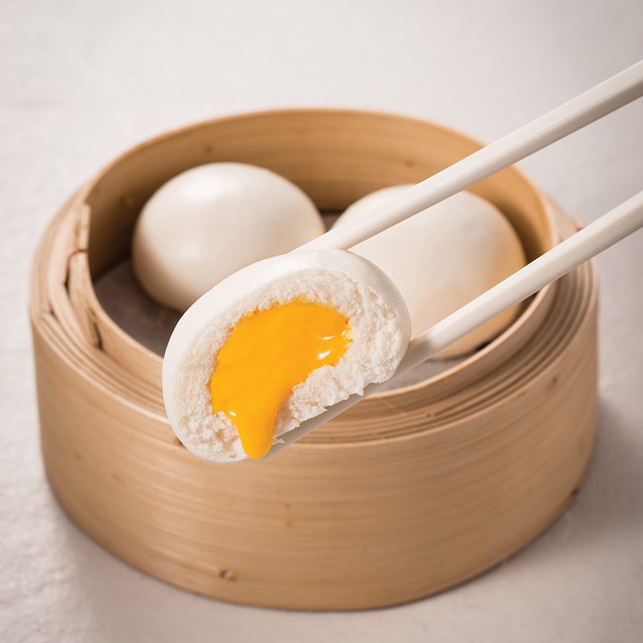 Chopsticks holding a steamed bun with a runny custard filling