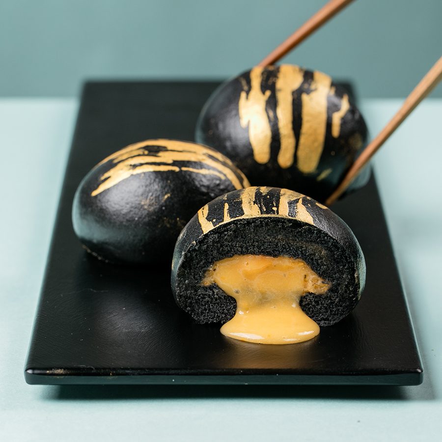 A plate of black steamed buns coated with edible gold flakes. One is sliced in half to reveal a runny custard filling