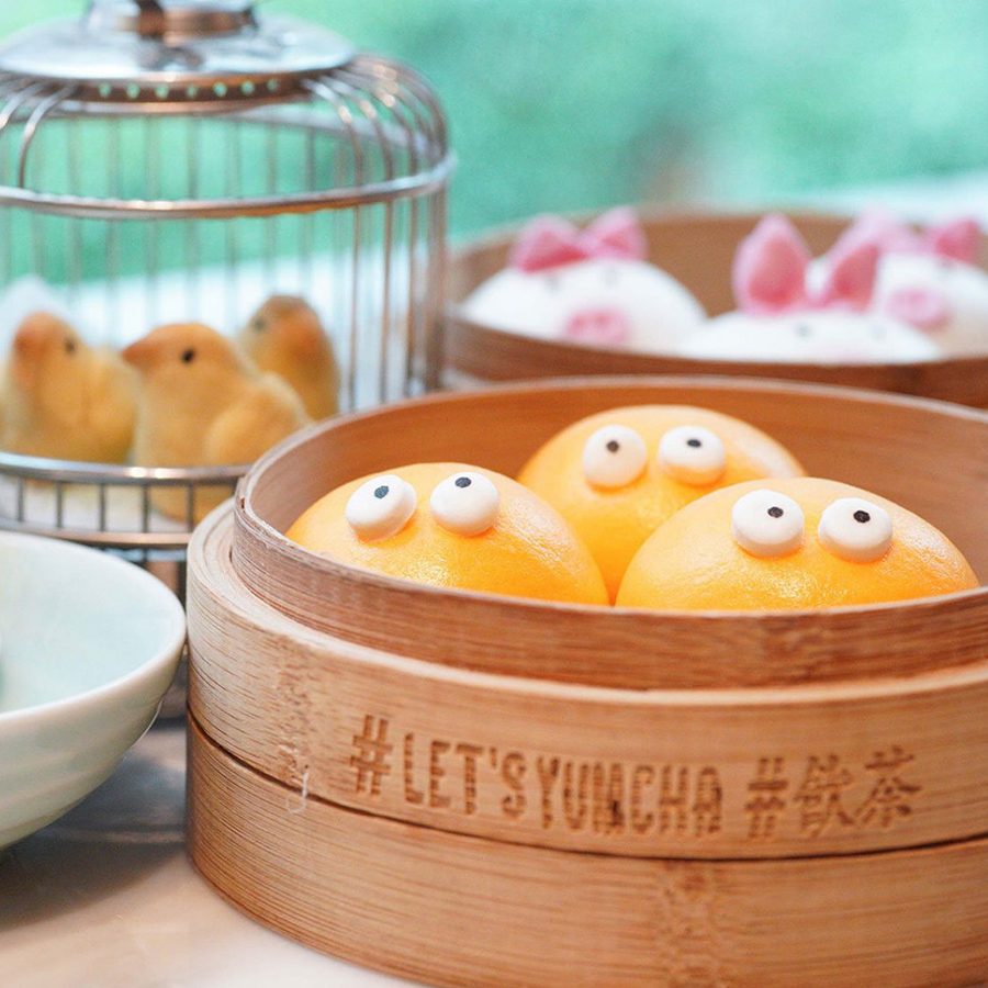 A basket of orange steamed buns with googly eyes. Behind it is a miniature bird cage containing bird-shaped pastries, and a basket of pink steamed buns. 
