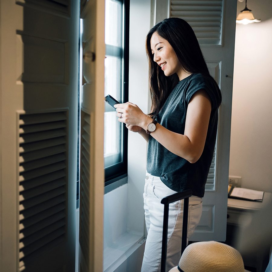 Asian female traveller standing by the window while checking her phone