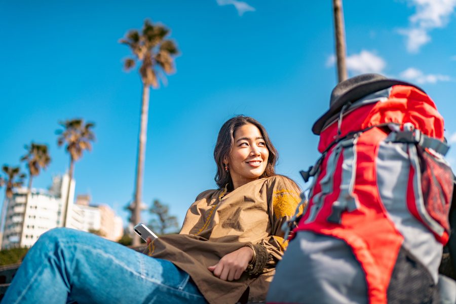Woman sitting on a bench underneath palm trees with a travel backpack 