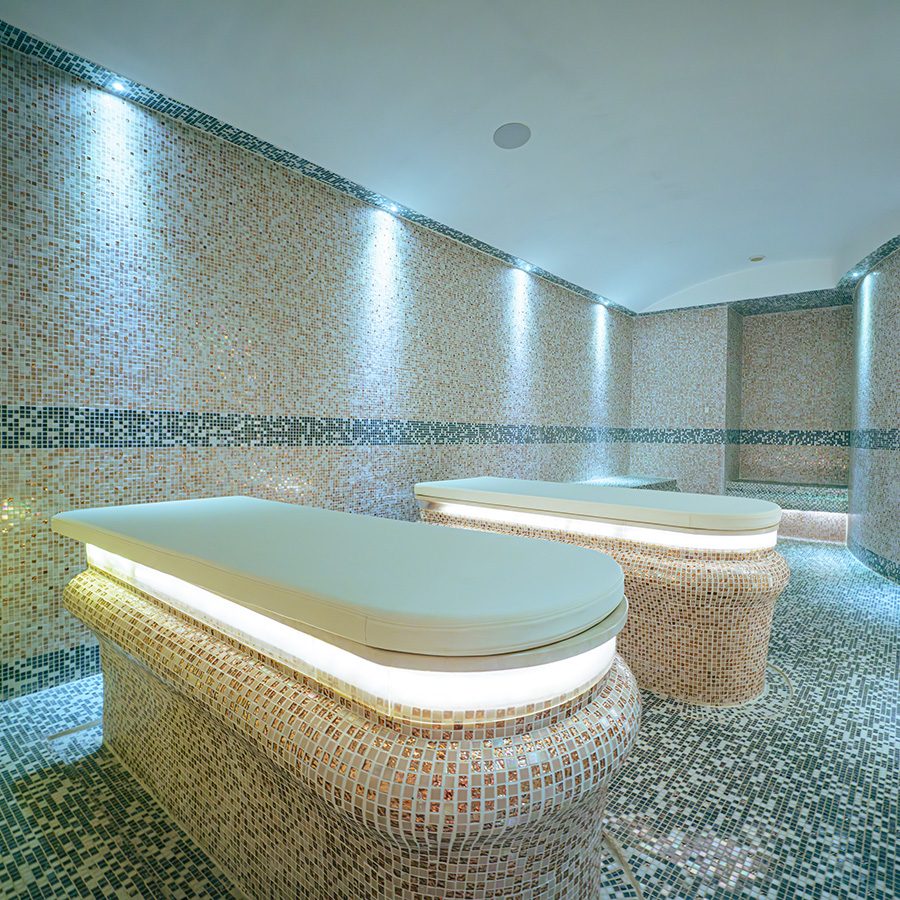 A room in Hui Spa with a tiled floor and walls, two lit-up beds, and a sink on the right