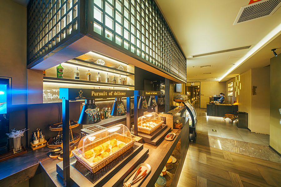 Covered trays of food on a table with tongs in front and beverage bottles displayed on shelves in Mi Spa’s relaxation area