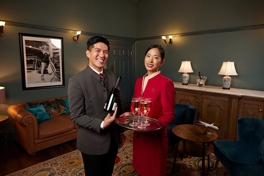 Two members of Cathay’s cabin crew, one man and one woman, hold a wine bottle and a tray of Champagne flutes inside the Cathay Suite at The London Palladium. The room has dark green walls with sconces, a brownish orange sofa, two deep blue chairs around a round wood table, and a patterned rug