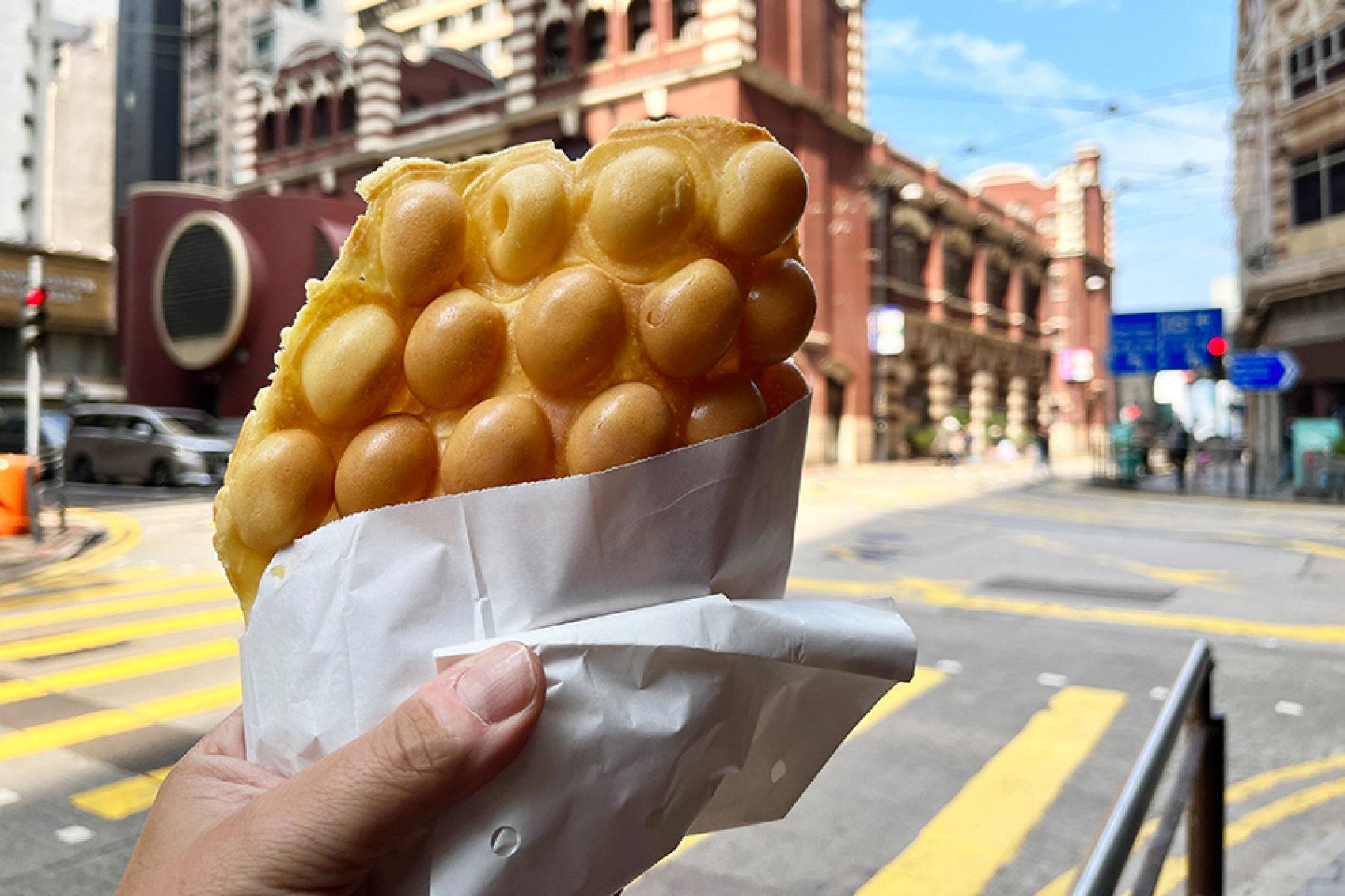 A traveller holds up a bag of egg waffles on the street in front of some buildings in Hong Kong. 