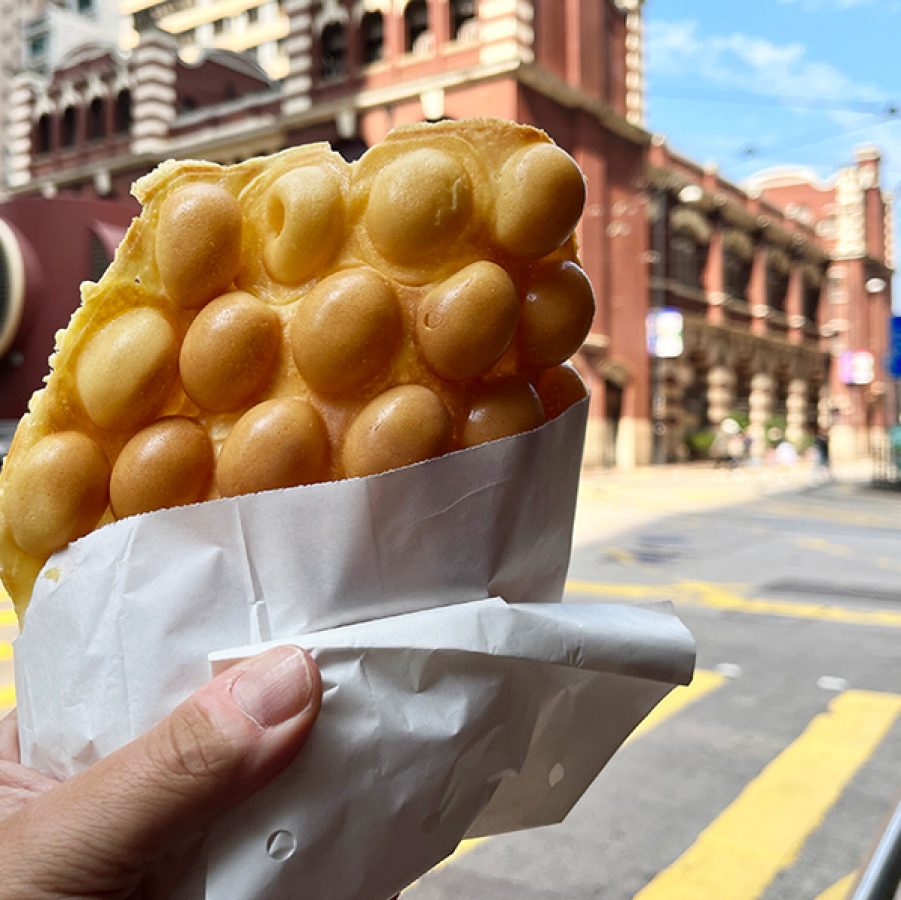 A traveller holds up a bag of egg waffles on the street in front of some buildings in Hong Kong. 