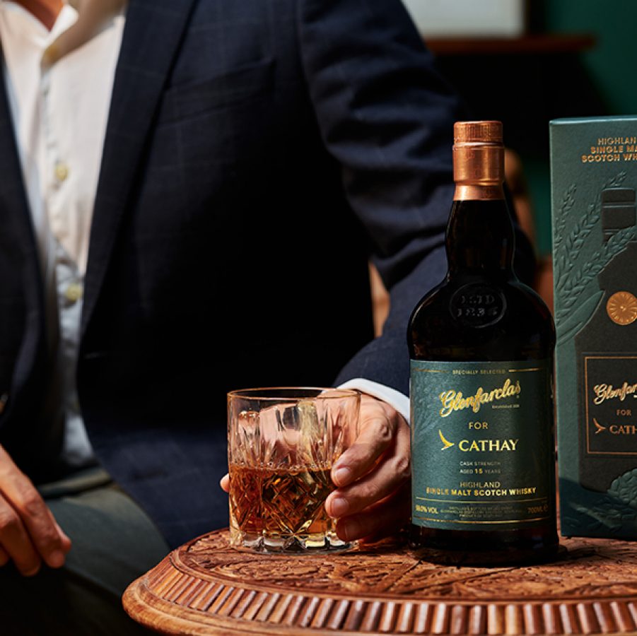 Cathay x Glenfarclas highland single malt whisky on a table beside the box it comes in and a tumbler of whiskey. A suited man is also sitting at the table.