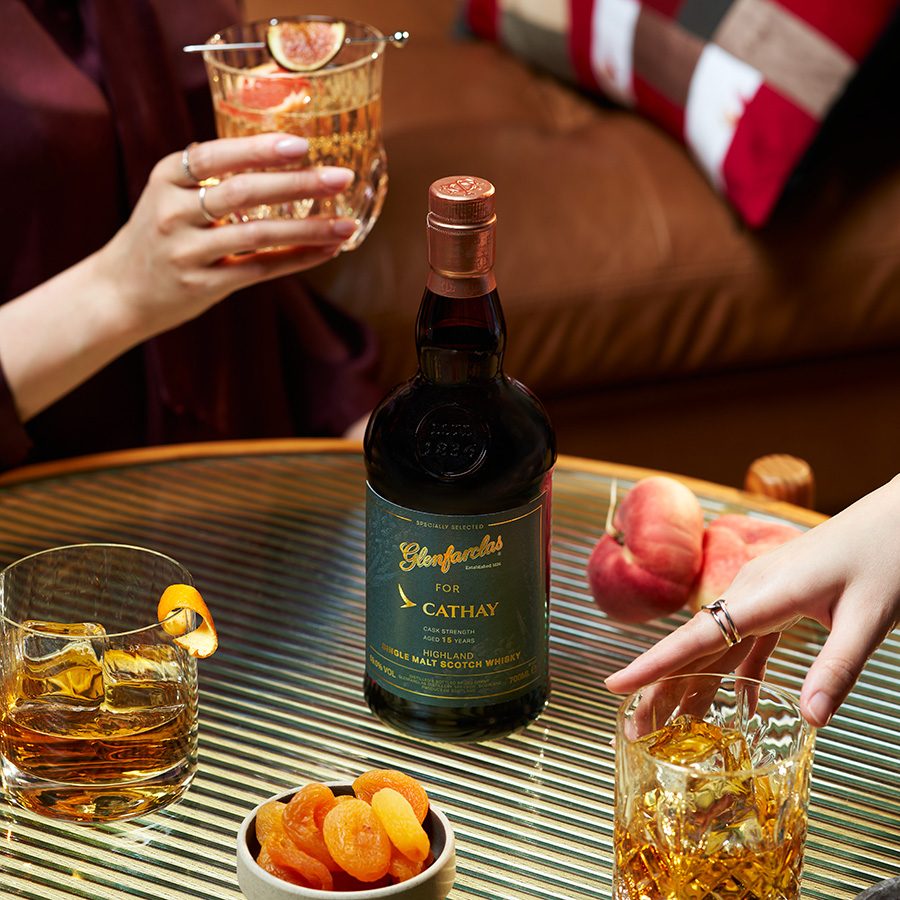 Shot of people drinking cocktails on a table featuring a Cathay plane, bottle of Cathay x Glenfarclas highland single malt whisky and fruits