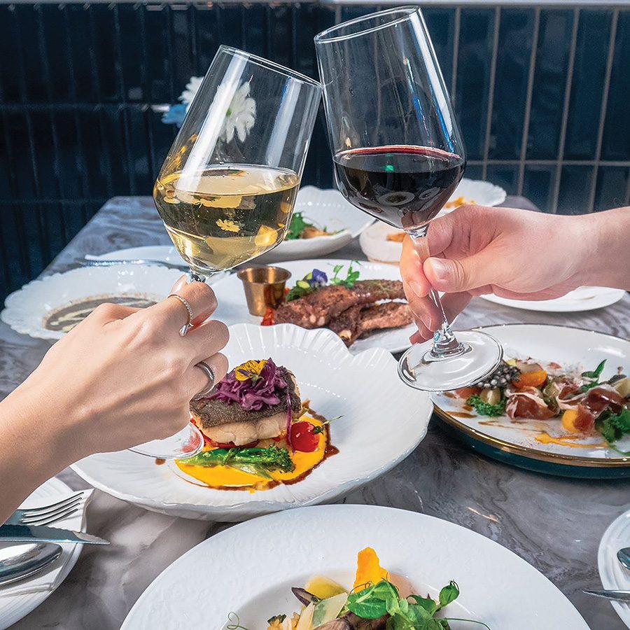 A pair of clinking wineglasses, one red one white, over a table of food at Royal Inn 