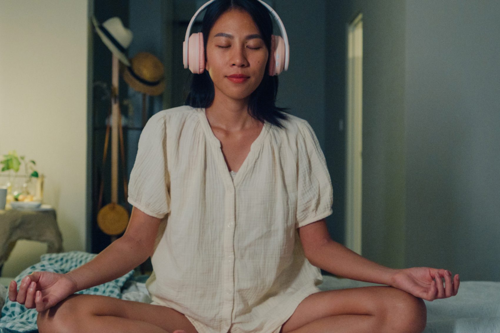 A woman sitting cross-legged on her bed with headphones on, eyes closed, and hands rested on her knees. Behind her, there is a lamp, plants, and a coat rack with hats. 