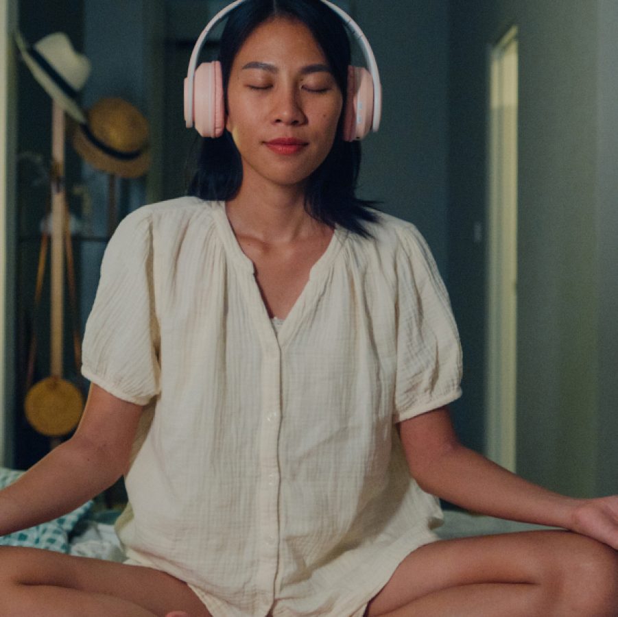 A woman sitting cross-legged on her bed with headphones on, eyes closed, and hands rested on her knees. Behind her, there is a lamp, plants, and a coat rack with hats. 