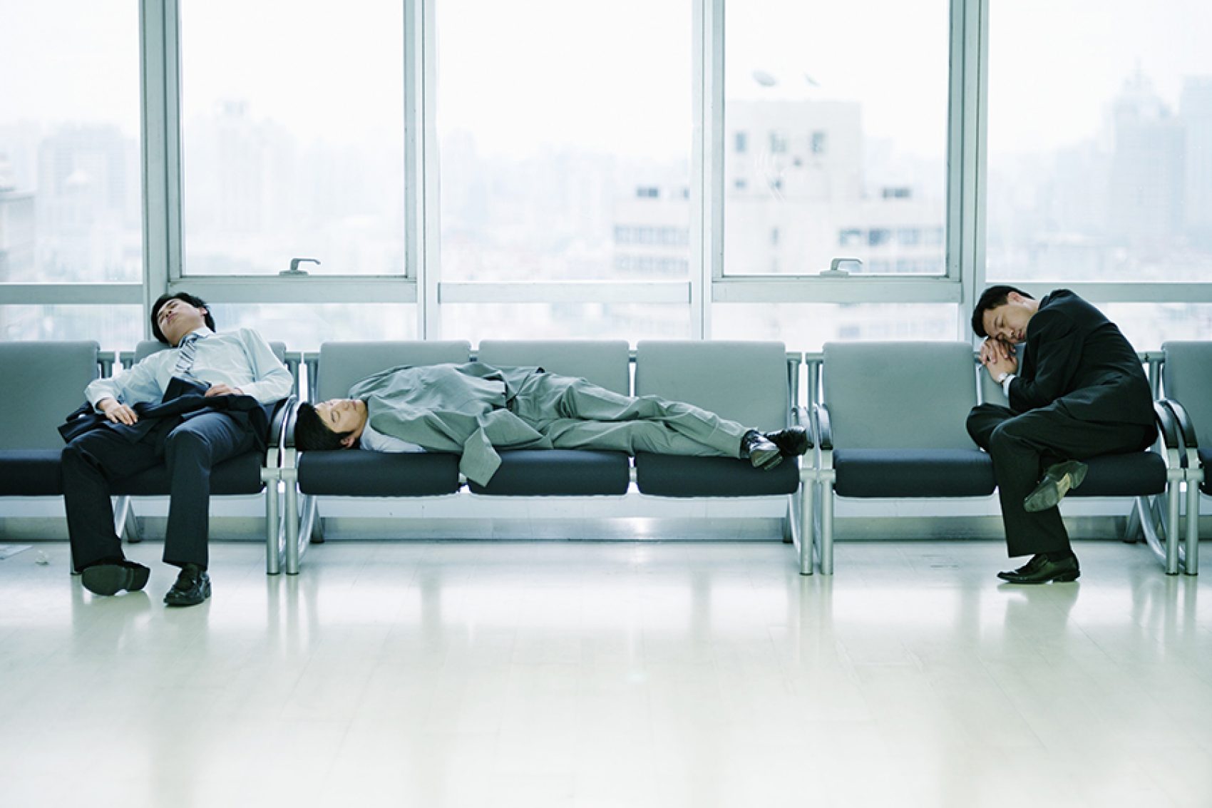 Three men dressed in business attire sleeping on chairs in an airport. 